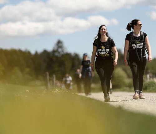 Im Vordergrund sind zwei schwarz gekleidete Frauen in Mammutmarsch T-Shirt zu sehen. Die eine lacht in die Kamera, die andere schaut zur Seite. Im Hintergrund folgen weitere Wanderer, die nur unscharf zu sehen sind.