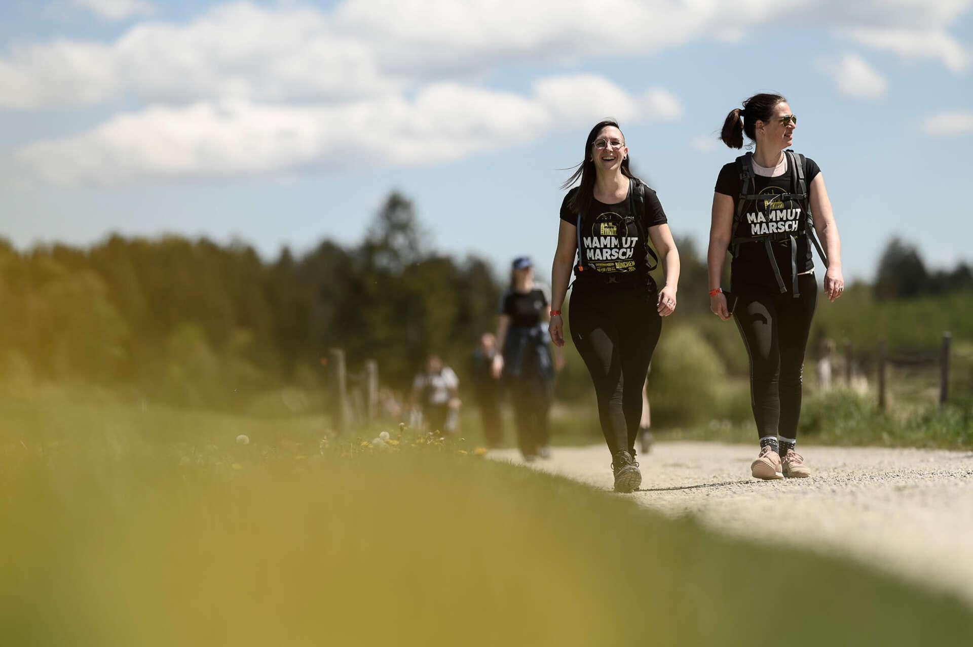 Im Vordergrund sind zwei schwarz gekleidete Frauen in Mammutmarsch T-Shirt zu sehen. Die eine lacht in die Kamera, die andere schaut zur Seite. Im Hintergrund folgen weitere Wanderer, die nur unscharf zu sehen sind.