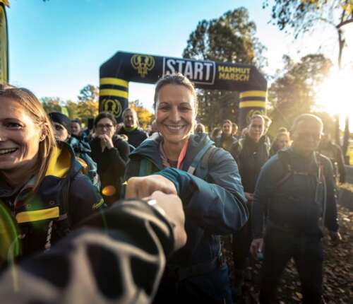 Teilnehmer des Mammutmarsch Stuttgart stehen motiviert am Startbogen und feiern den Beginn der Herausforderung bei Sonnenaufgang, bereit für die lange Wanderung. Motivierender Abklatsch.