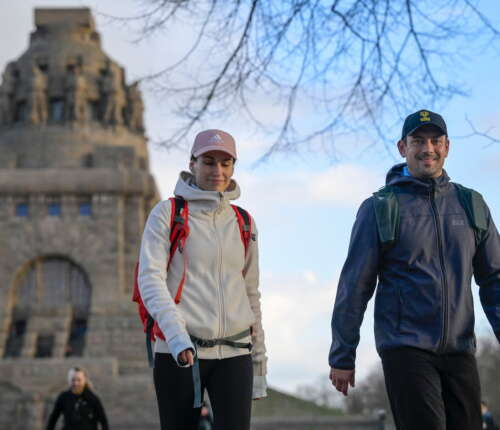 Zwei Personen in Wanderkleidung vor einem historischen Monument mit Rucksäcken und Mützen für eine Wanderung gerüstet