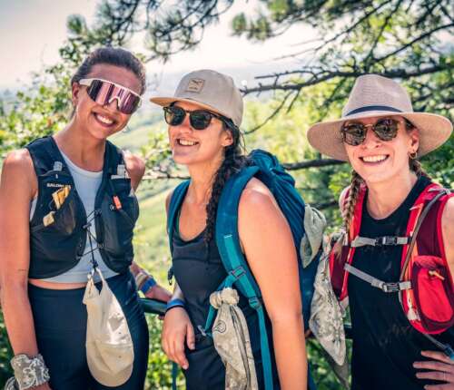 Drei Frauen in moderner Wanderkleidung, ausgestattet mit Rucksäcken und Hüten, genießen eine sonnige Wanderung in der Natur.