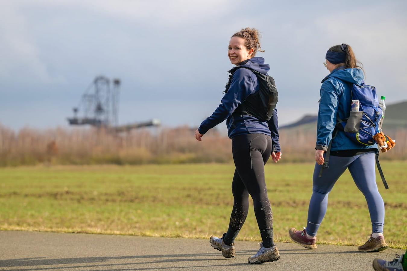 Zwei Frauen in Wanderkleidung mit Rucksäcken, die auf einer Landstraße entlanggehen, eine davon lächelt in die Kamera, im Hintergrund ist eine industrielle Anlage zu sehen.