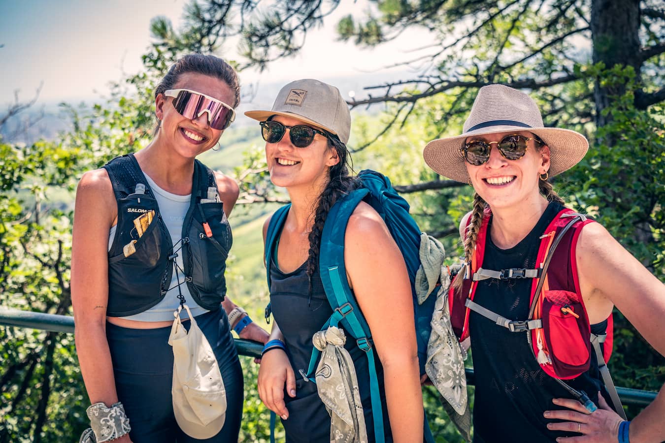 Drei Frauen in moderner Wanderkleidung, ausgestattet mit Rucksäcken und Hüten, genießen eine sonnige Wanderung in der Natur.