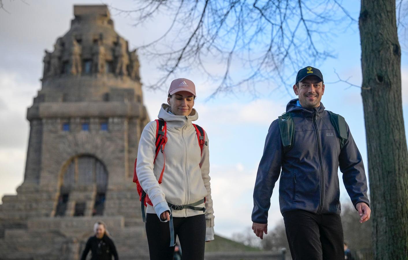 Zwei Personen in Wanderkleidung vor einem historischen Monument mit Rucksäcken und Mützen für eine Wanderung gerüstet