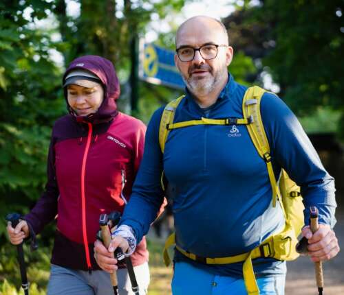 Mann und Frau in hochwertigen Wanderklamotten und Wanderstöcken. Er ist blau gekleidet mit einem gelben Rucksack. Sie trägt eine rot-lila Hardshelljacke.
