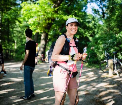 Lächelnde, sportliche, rosa gekleidete Frau mit Cap, Wanderrucksack, pinken Trailrunning-Stöcken und Sonnenbrille in den Händen