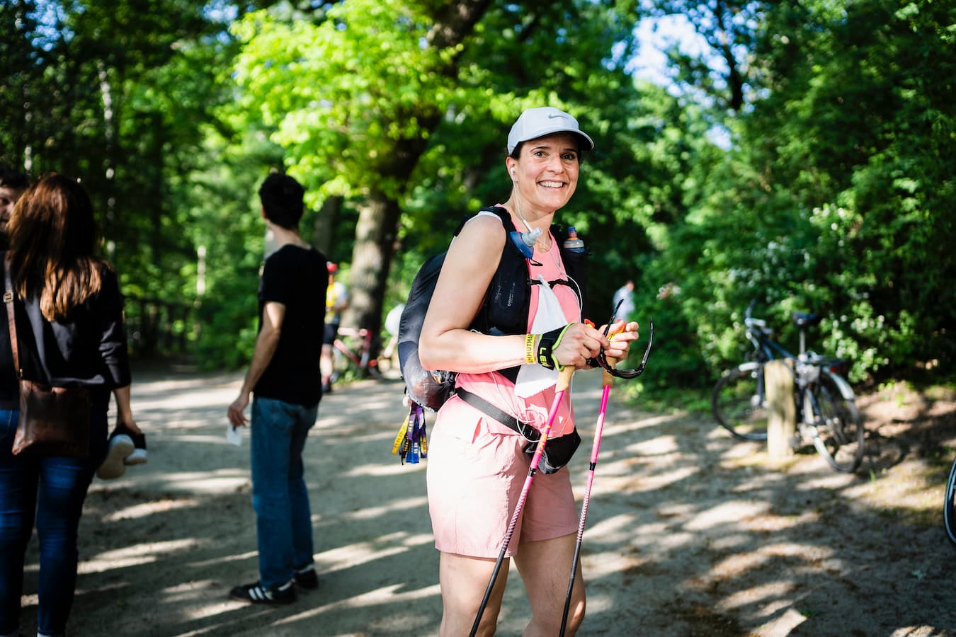 Lächelnde, sportliche, rosa gekleidete Frau mit Cap, Wanderrucksack, pinken Trailrunning-Stöcken und Sonnenbrille in den Händen