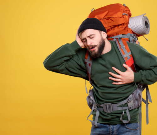 In grünem Pulli gekleideter Mann mit großem Rucksack auf dem Rücken hält sich angestrengt den Kopf und fasst sich mit der anderen Hand ans Herz