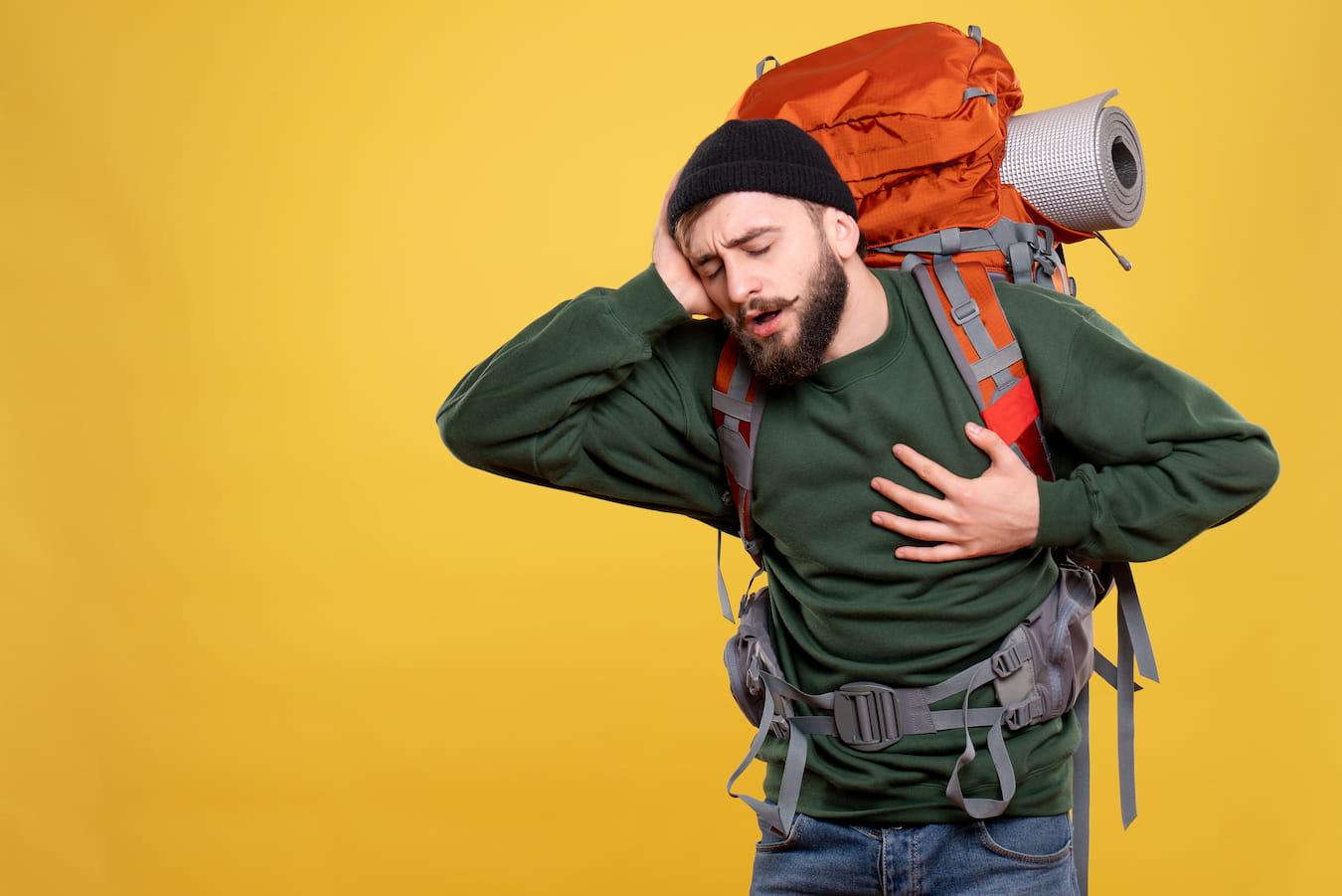 In grünem Pulli gekleideter Mann mit großem Rucksack auf dem Rücken hält sich angestrengt den Kopf und fasst sich mit der anderen Hand ans Herz