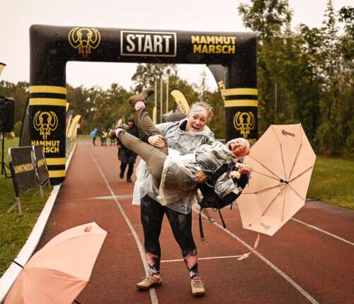 Zwei lachende Frauen in Regenponchos beim Mammutmarsch Ziel. Die eine hebt die andere, die einen Schirm in der Hand hat.