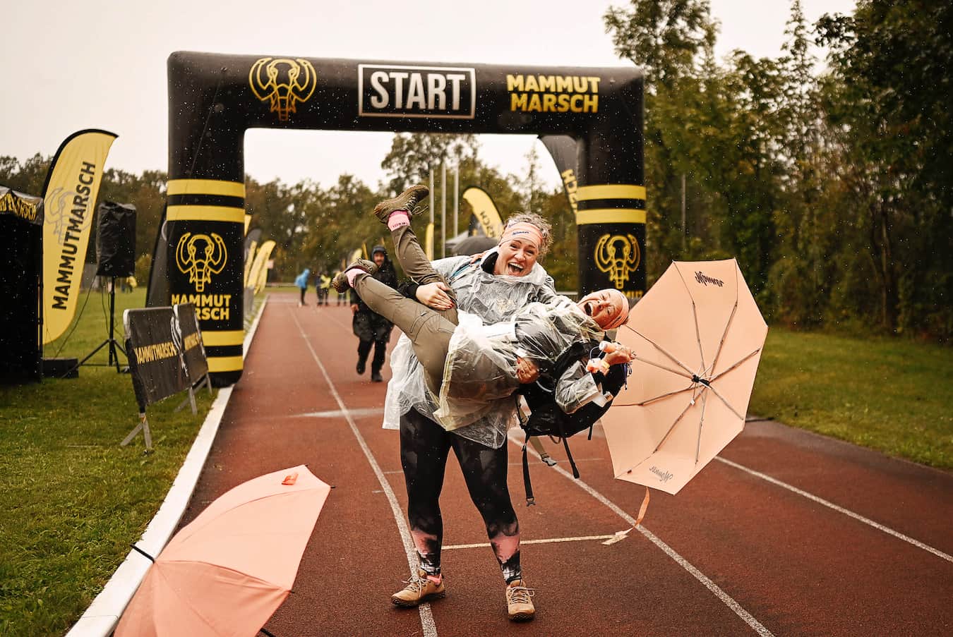 Zwei lachende Frauen in Regenponchos beim Mammutmarsch Ziel. Die eine hebt die andere, die einen Schirm in der Hand hat.