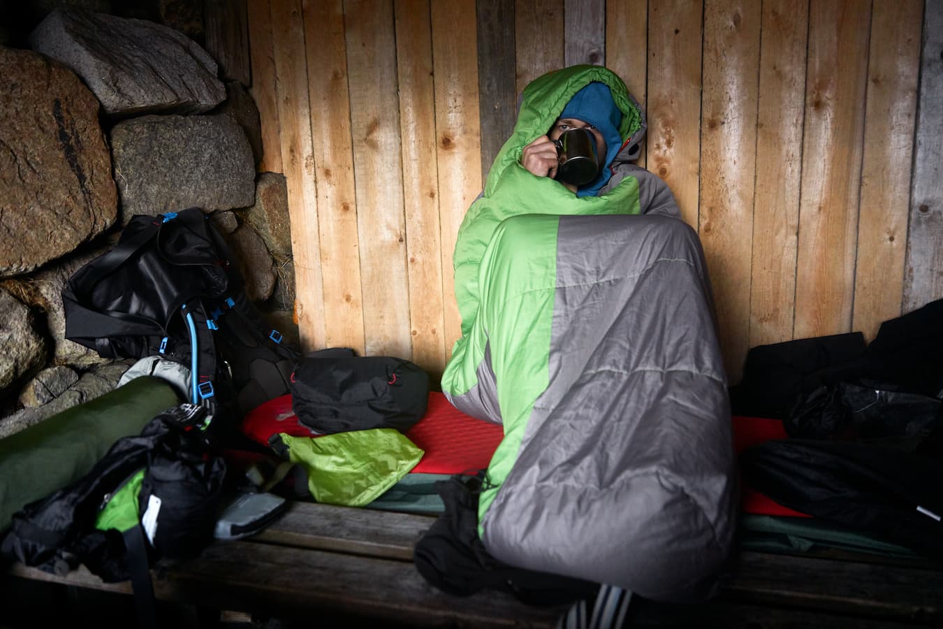 Mann sitzt eingemummelt in grau-grünem Schlafsack und trinkt aus einem Becher