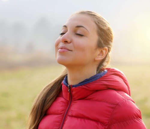 Junge Frau mit Pferdeschwanz und in pinker Daunenjacke hat die Augen geschlossen und atmet genüsslich durch die Nase ein.