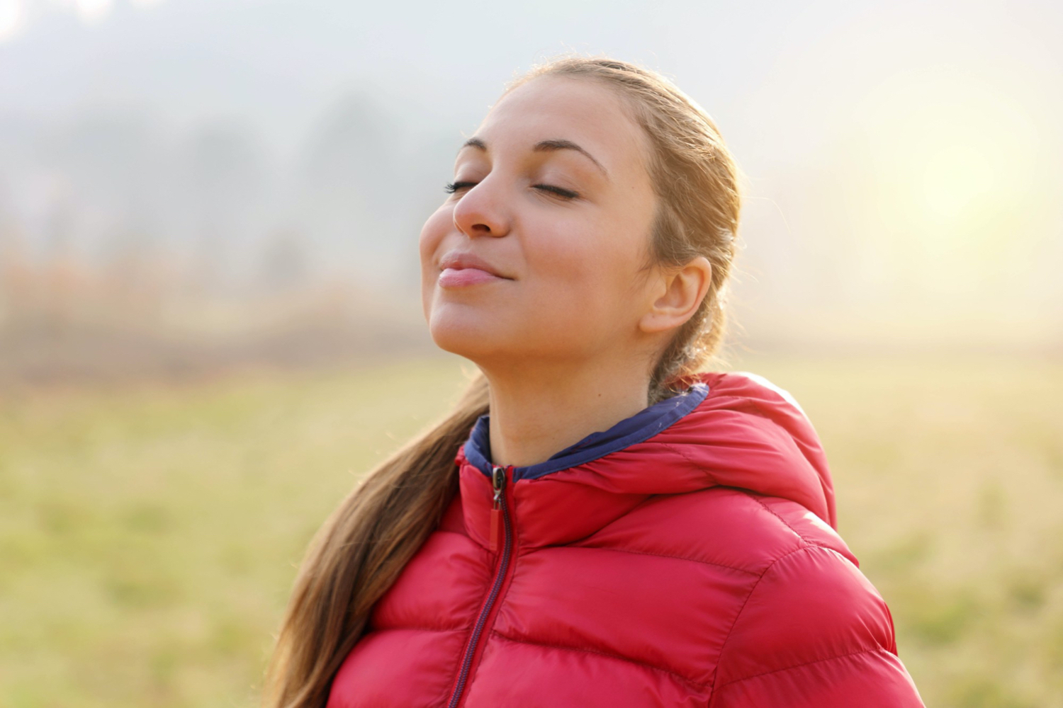 Junge Frau mit Pferdeschwanz und in pinker Daunenjacke hat die Augen geschlossen und atmet genüsslich durch die Nase ein.