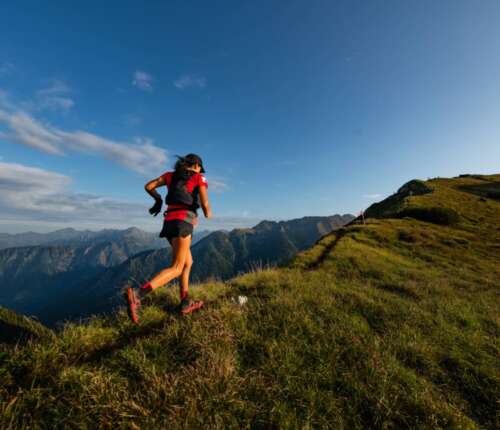 Eine Person in einem roten Oberteil und Shorts läuft unter blauem Himmel einen grasbewachsenen Bergrücken hinauf, im Hintergrund sind in der Ferne Berge zu sehen.