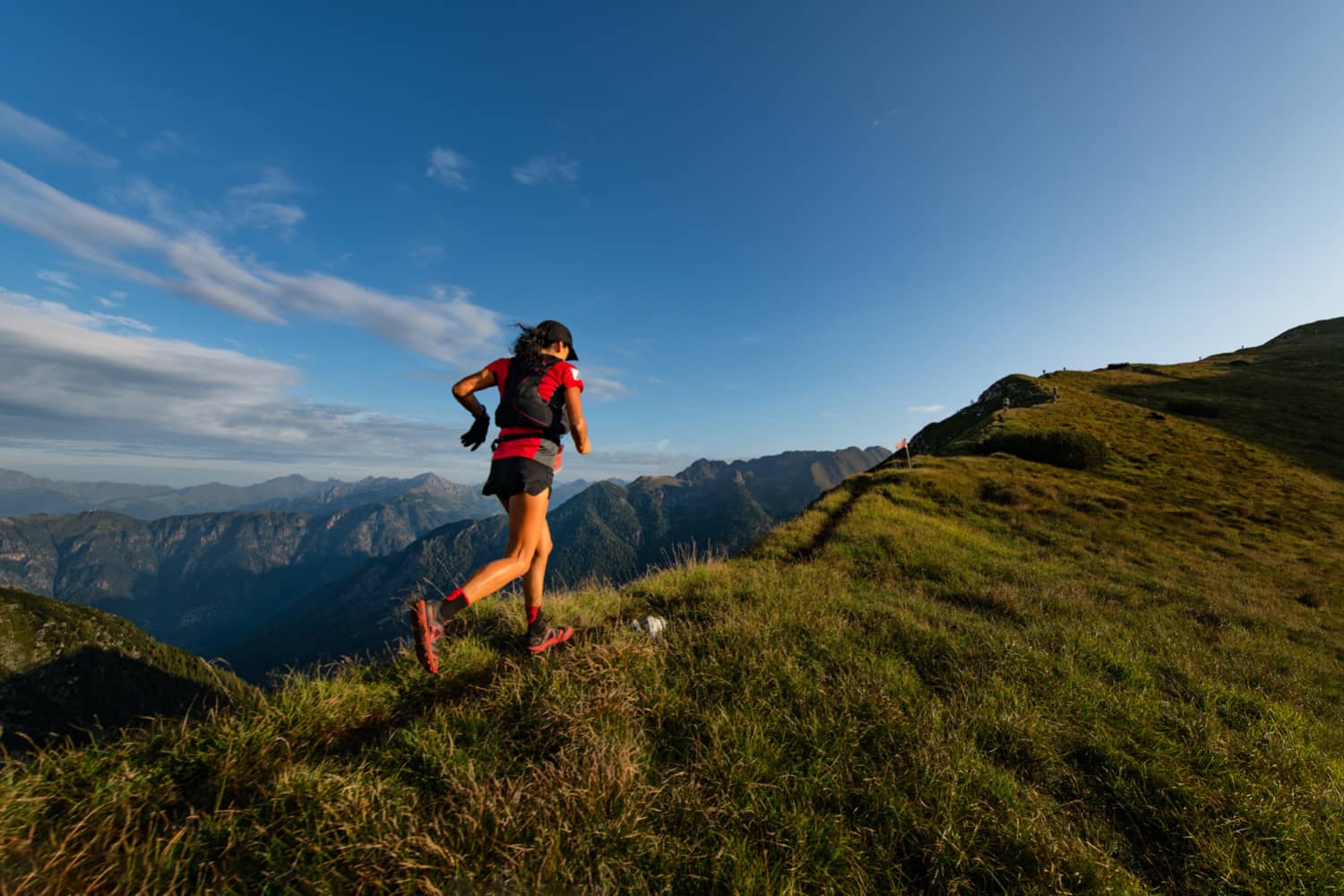Eine Person in einem roten Oberteil und Shorts läuft unter blauem Himmel einen grasbewachsenen Bergrücken hinauf, im Hintergrund sind in der Ferne Berge zu sehen.