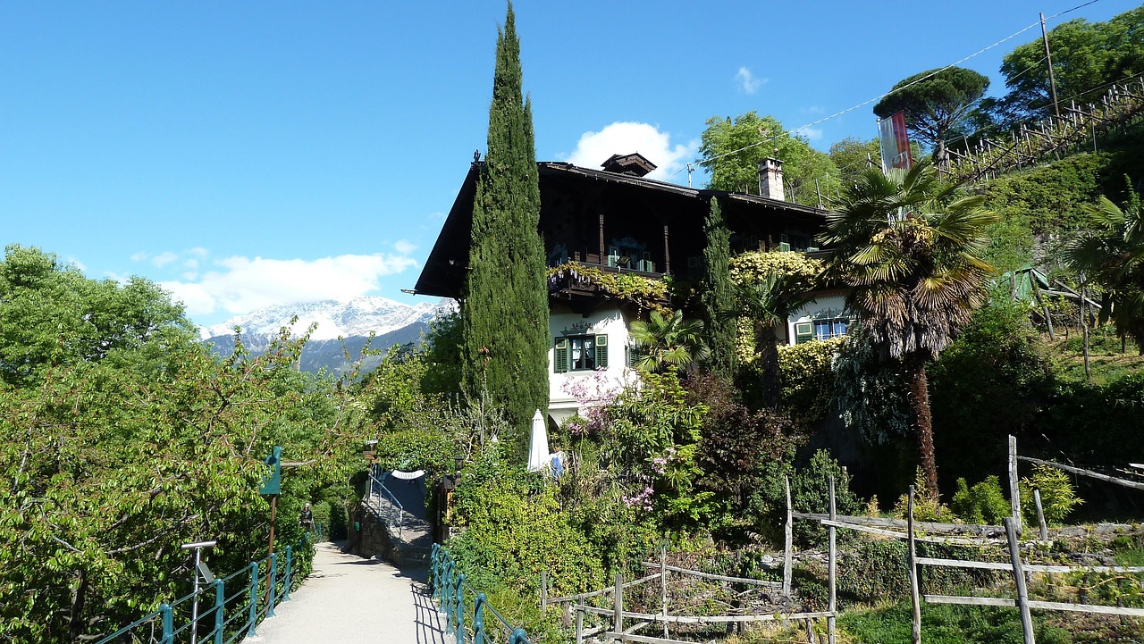 Ein schmaler Wanderweg führt an einem traditionellen Haus mit üppiger Vegetation vorbei, im Hintergrund ragen schneebedeckte Berge auf.