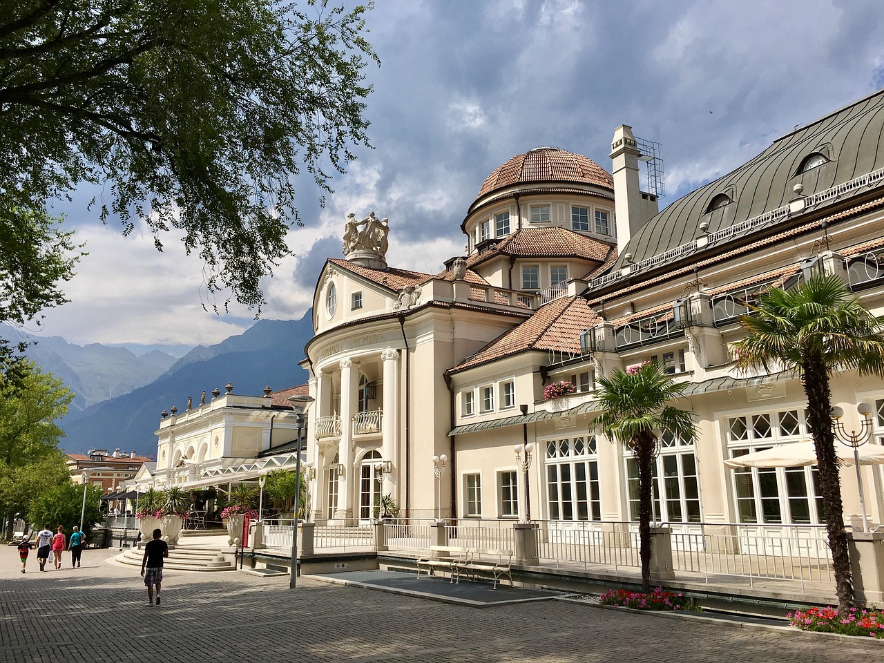 Ein prachtvolles historisches Kurhaus mit Kuppeldach und Säulen steht an einer Promenade, im Hintergrund die Berge.