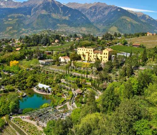 Blick auf Schloss Trauttmansdorff in Meran, eingebettet in die üppig grüne Landschaft der Gärten mit Teich, Terrassen und den Südtiroler Bergen im Hintergrund.
