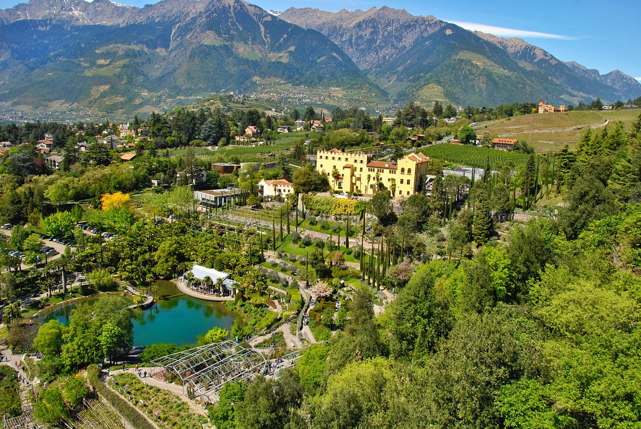 Blick auf Schloss Trauttmansdorff in Meran, eingebettet in die üppig grüne Landschaft der Gärten mit Teich, Terrassen und den Südtiroler Bergen im Hintergrund.