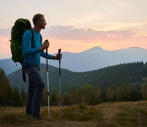 Ein Mann beim Wandern, er steht mit Wanderrucksack und Wanderstöcken auf einer Anhöhe und bewundert den Ausblick.