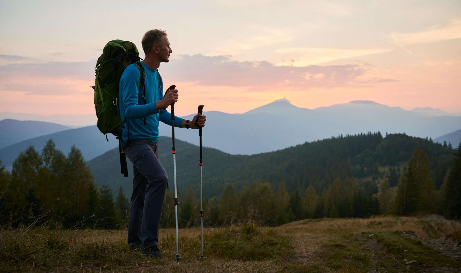Ein Mann beim Wandern, er steht mit Wanderrucksack und Wanderstöcken auf einer Anhöhe und bewundert den Ausblick.